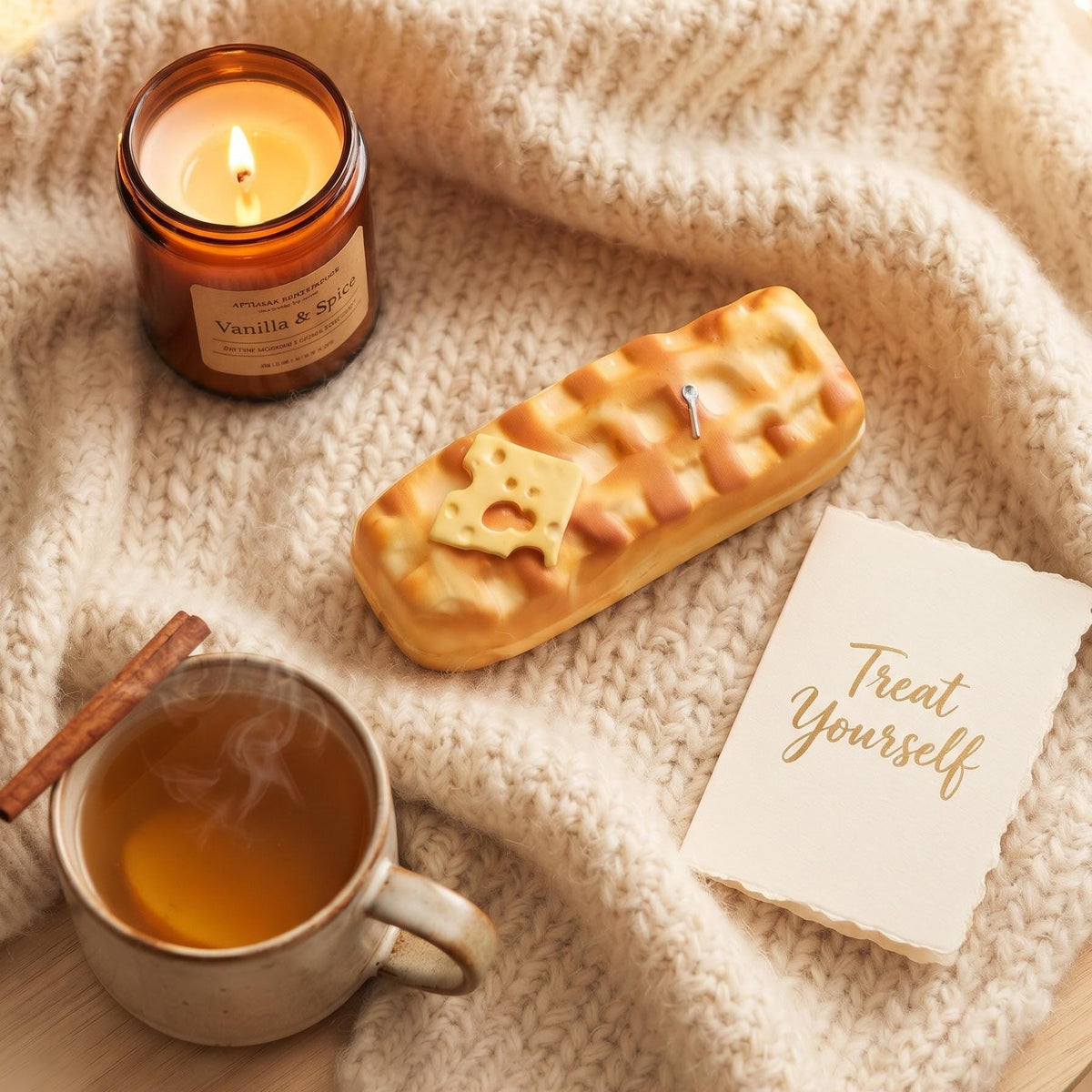 A cozy flat-lay scene with a lit amber glass candle labeled Vanilla & Spice, a knitted cream blanket, a small rectangular pastry with a bear-shaped cookie on top, a mug of hot tea with a cinnamon stick, and a note that says 'Treat Yourself'.