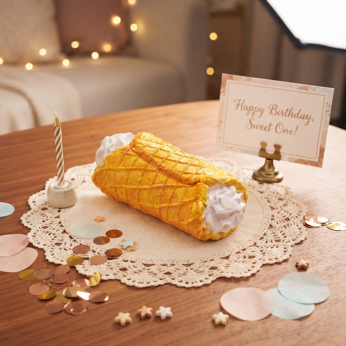 A decorative birthday cake prop shaped like a waffle cone with white whipped frosting ends sits on a lace doily on a wooden table. A small candle, a card reading 'Happy Birthday, Sweet One!', and scattered pastel confetti are arranged around it in a softly lit indoor setting.