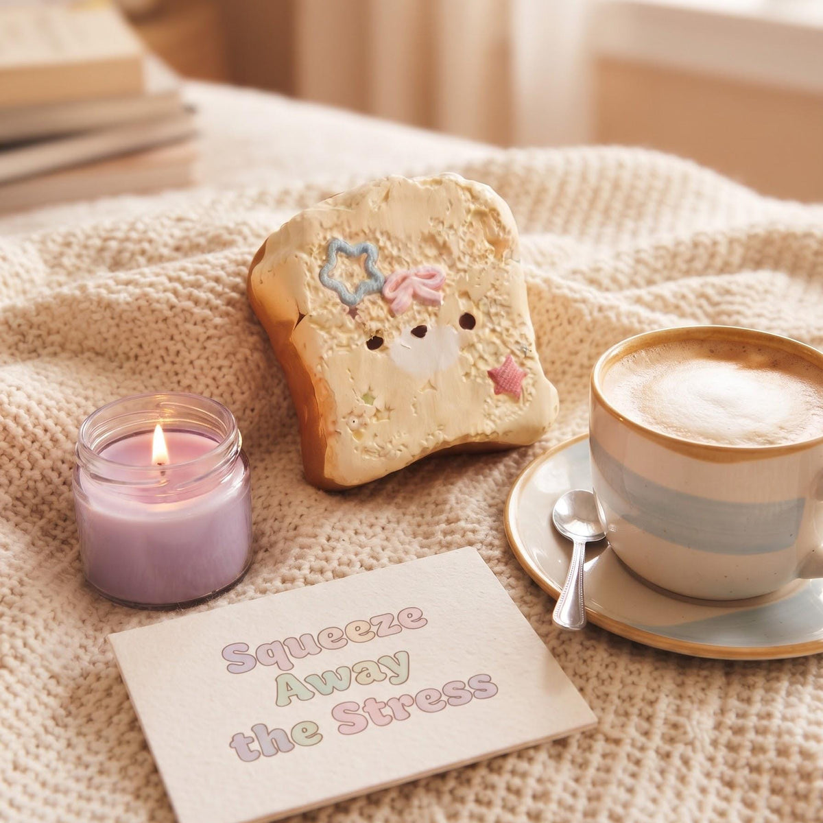 A cozy lifestyle scene on a cream knit blanket featuring a slice of decorated toast with a cute bear face, a purple candle, a cup of cappuccino on a saucer with a spoon, and a card that reads 'Squeeze Away the Stress'.