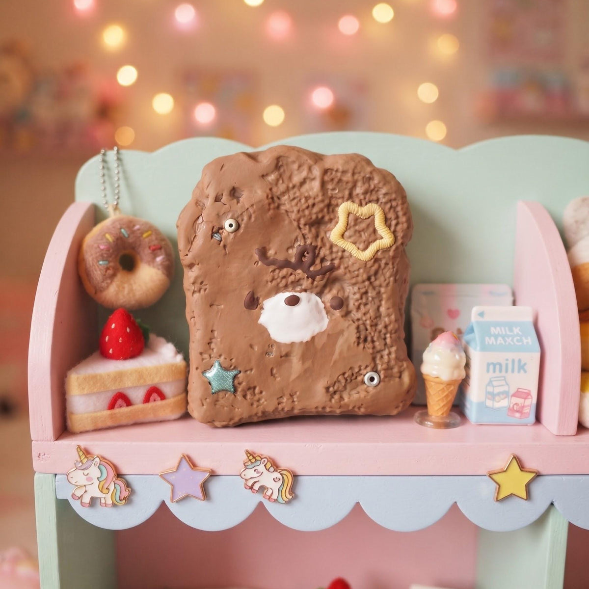 A cute, pastel-colored toy playset featuring a central brown bread/toast character with a bear face, surrounded by miniature donuts, cakes, an ice cream cone, and a milk carton, arranged on a pink and mint green shelf against a warm bokeh background with string lights.