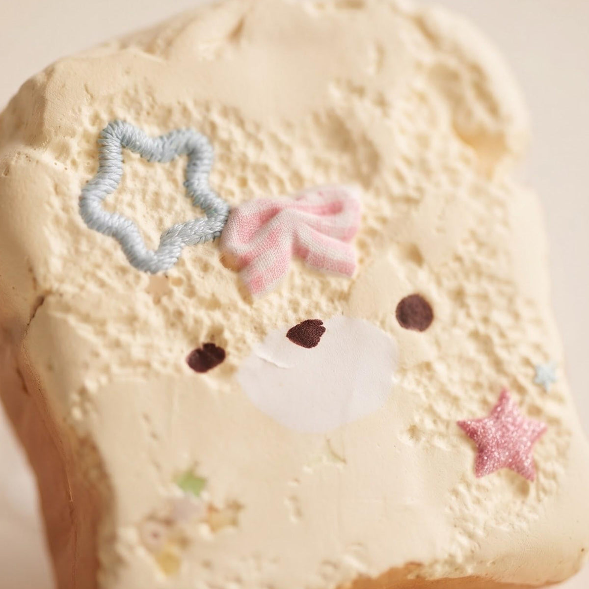 A cute, bear-face decorated slice of toast or cookie with cream-colored frosting, featuring a pink bow, blue star, and pink glitter star on a warm beige background.