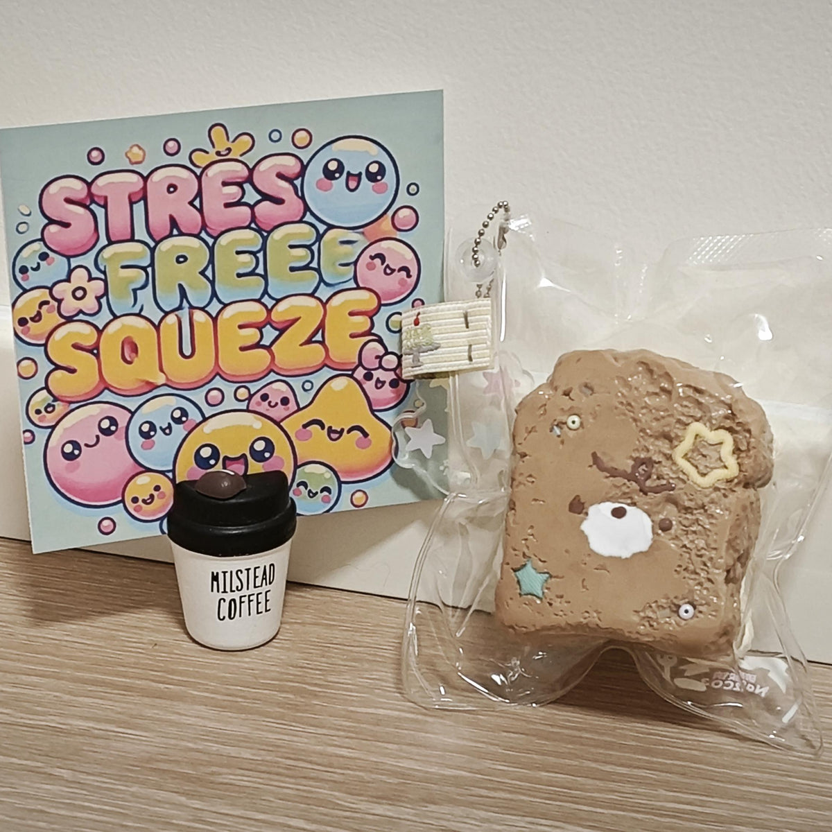 A cute tan bread-slice-shaped squishy toy with a bear face and star details, shown in a clear plastic bag next to a small black coffee cup charm and a colorful 'Stress Free Squeeze' card on a light wood floor.