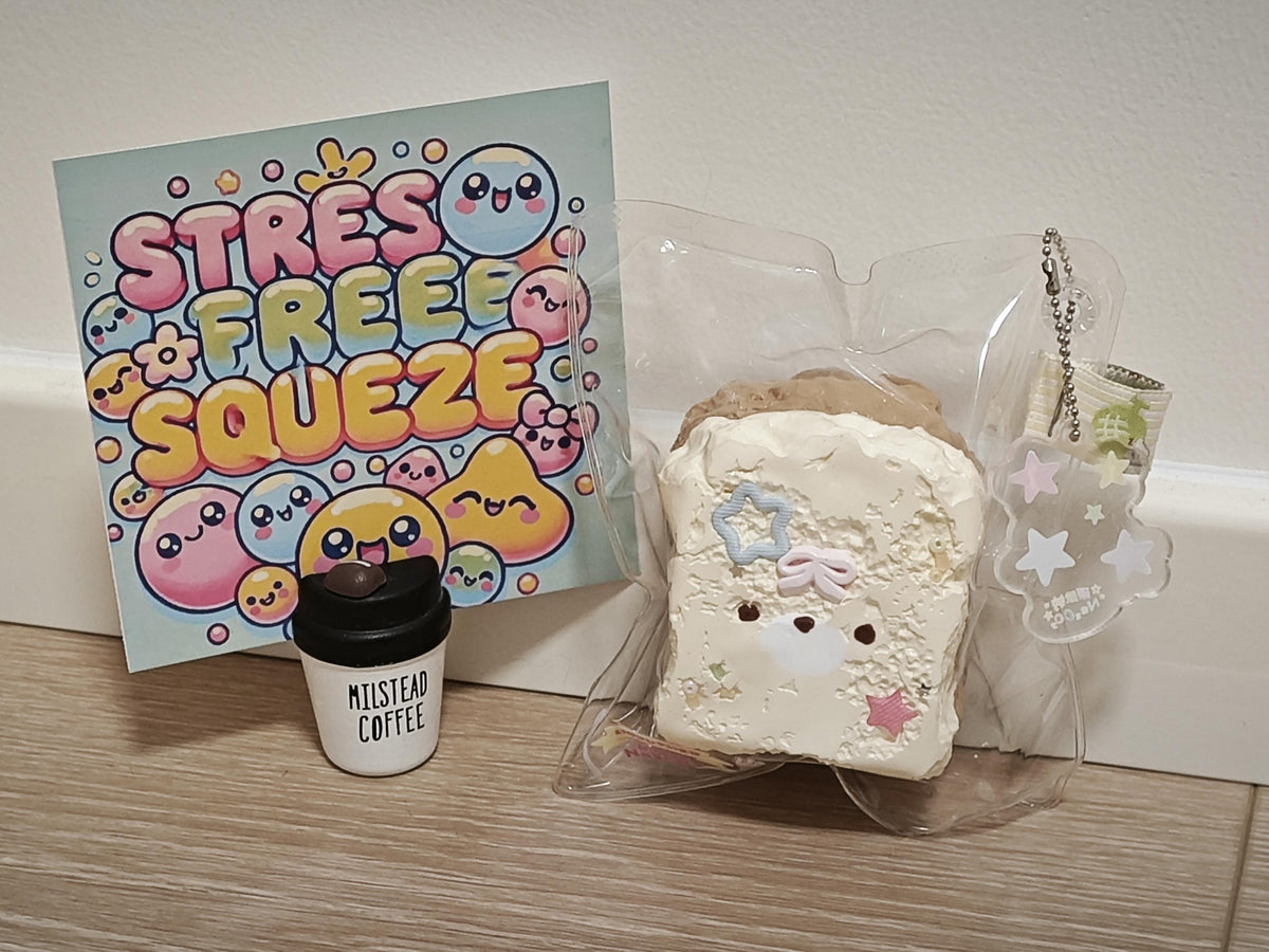 A cute, pastel-colored 'Stress Free Squeeze' plush toy shaped like a slice of toast with a smiling face, shown in its clear plastic packaging next to a small cup labeled 'Milstead Coffee' and a colorful 'Stress Free Squeeze' sign.
