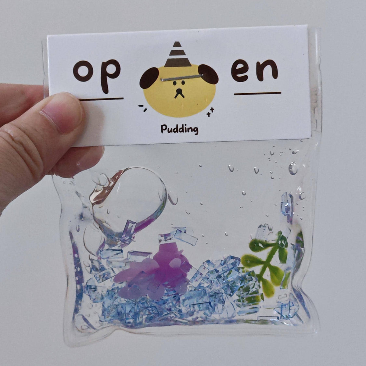 A hand holds a clear plastic pouch with a white paper header card that reads 'open Pudding' with a cartoon dog face. Inside the pouch is a clear gel or liquid containing blue, purple, and green decorative pieces.