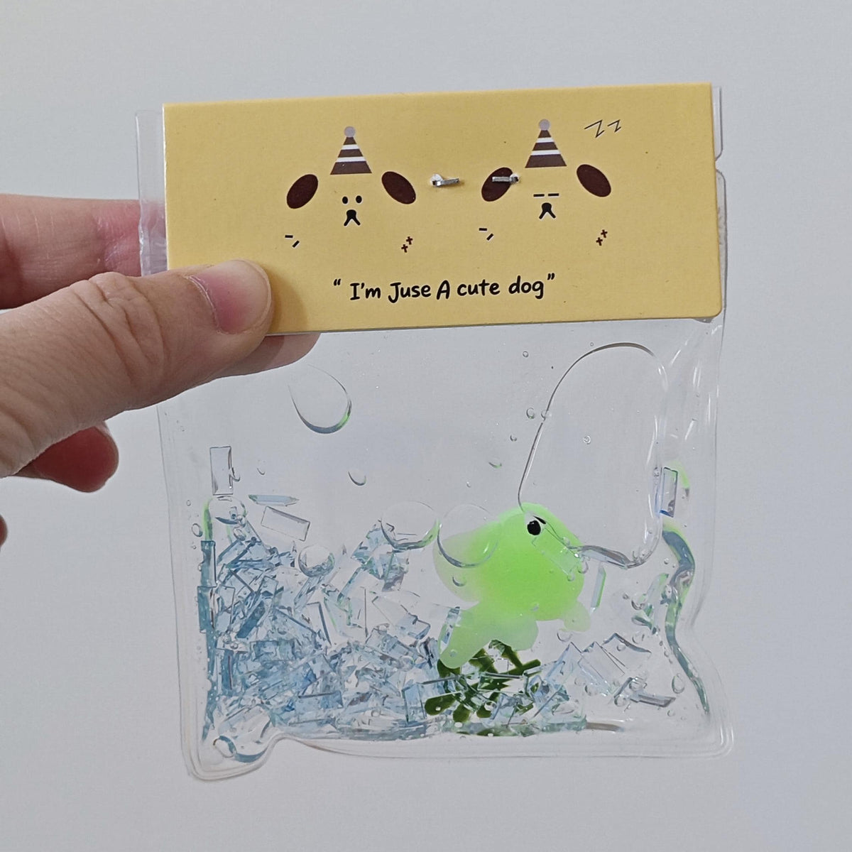 A hand holding a clear plastic bag containing a small green dog-shaped toy surrounded by blue and clear bubble-like pieces, with a yellow paper header card that reads "I'm Juse A cute dog" and shows cartoon dogs wearing party hats.