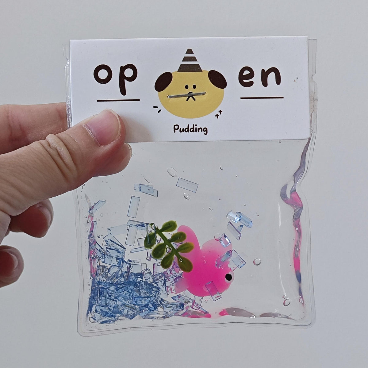 A hand holds a clear plastic pouch with a white paper flap that reads 'op en Pudding' with a cartoon face. Inside the pouch is a pink, squishy, novelty toy filled with colorful confetti and small clear pieces.