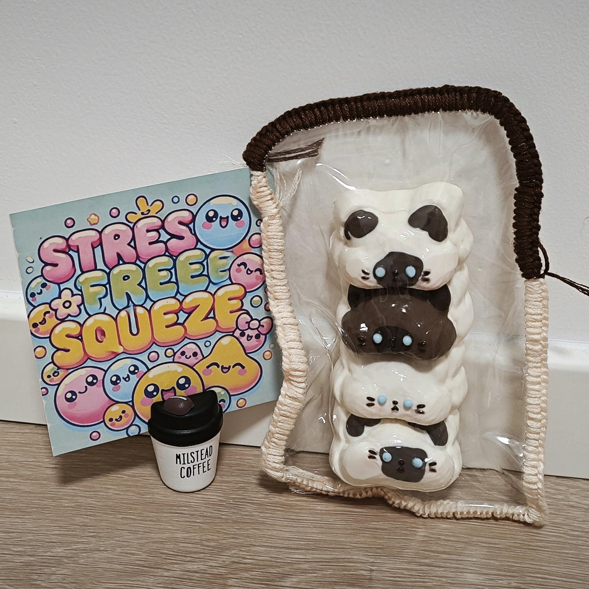 A stack of four cute cat-shaped squishy toys in white and dark brown, packaged in a clear zippered pouch with a cream cord. A small coffee cup accessory labeled 'MISTLETOE COFFEE' and a colorful 'STRESS FREE ZQUE' card are placed beside it on a wooden surface against a white wall.