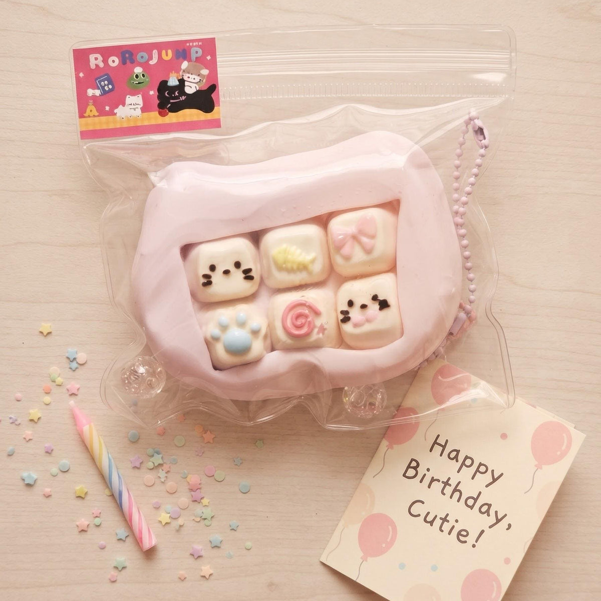 A pink silicone phone case with six cute character candy windows, packaged in a clear zip bag with a 'Roneijou' card and a 'Happy Birthday, Cutie!' card on a light wood background.