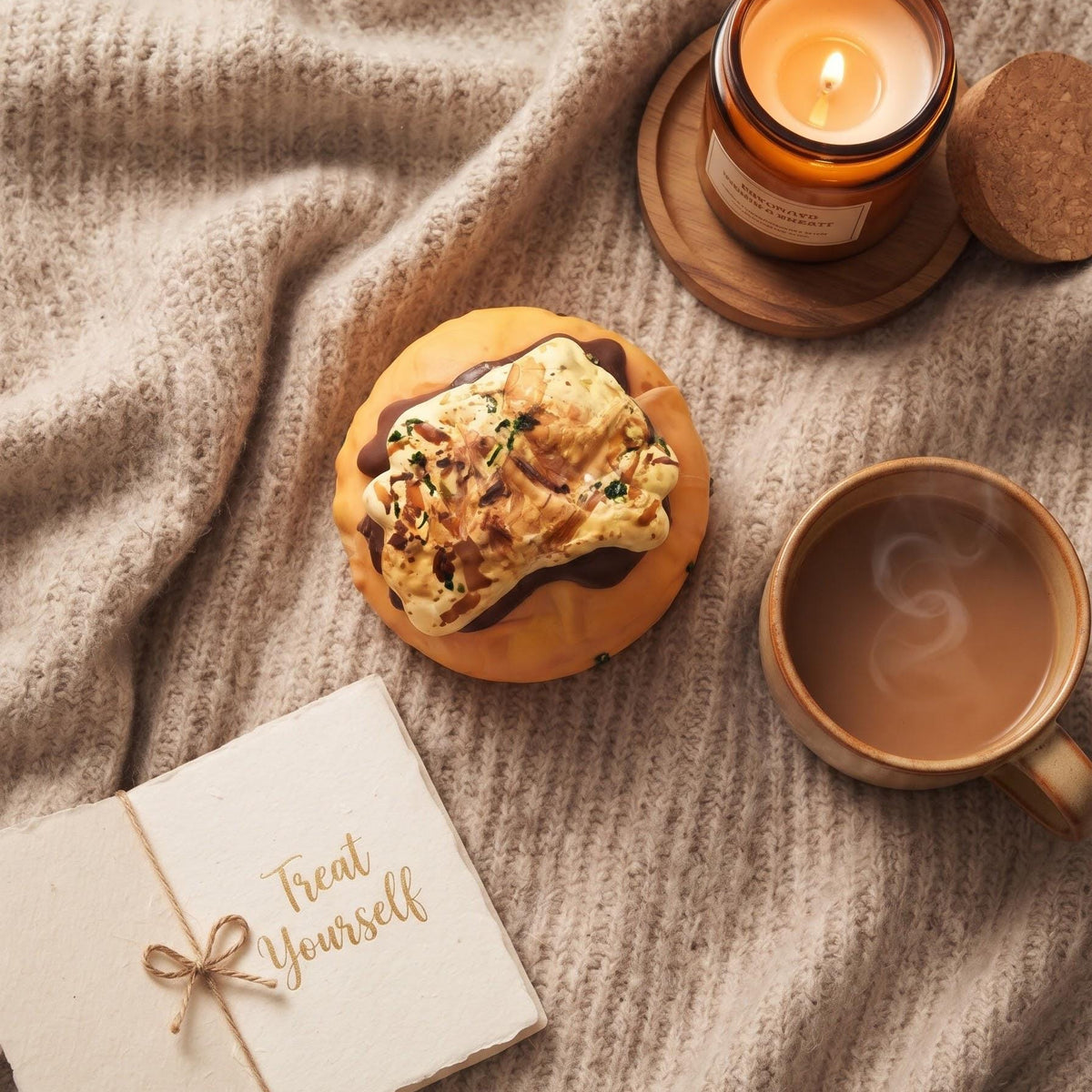 A cozy flat-lay scene on a beige knit blanket with a chocolate chip cookie topped with cream and nuts, a lit amber candle on a wooden coaster, a cup of coffee, and a small card that says 'Treat Yourself'.