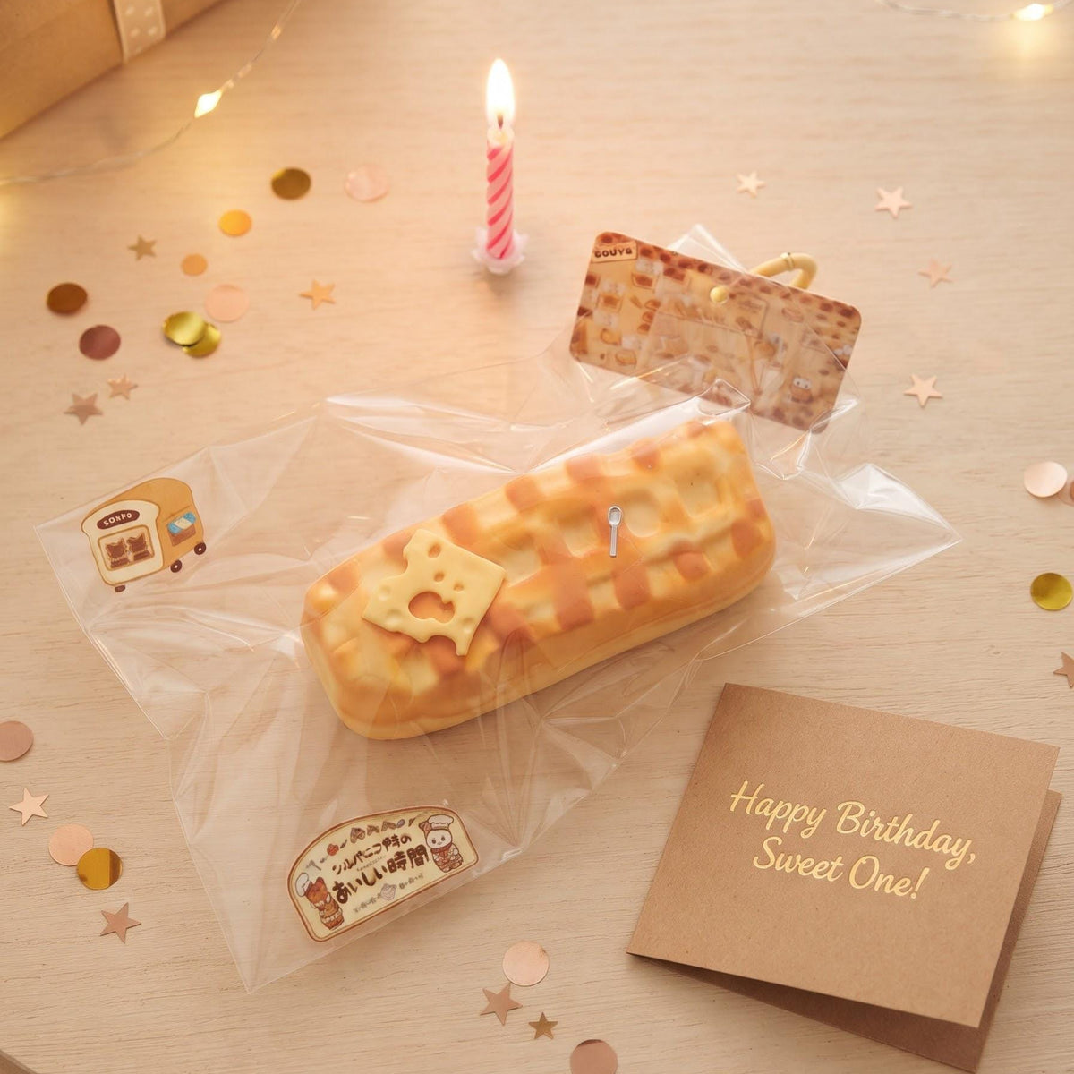 A packaged baked good or novelty food item sits on a light wooden table with warm string lights, confetti, a candle, and a brown card that reads 'Happy Birthday, Sweet One!'. The main product appears to be a long pastry or cake-like item in clear plastic packaging with printed labels.