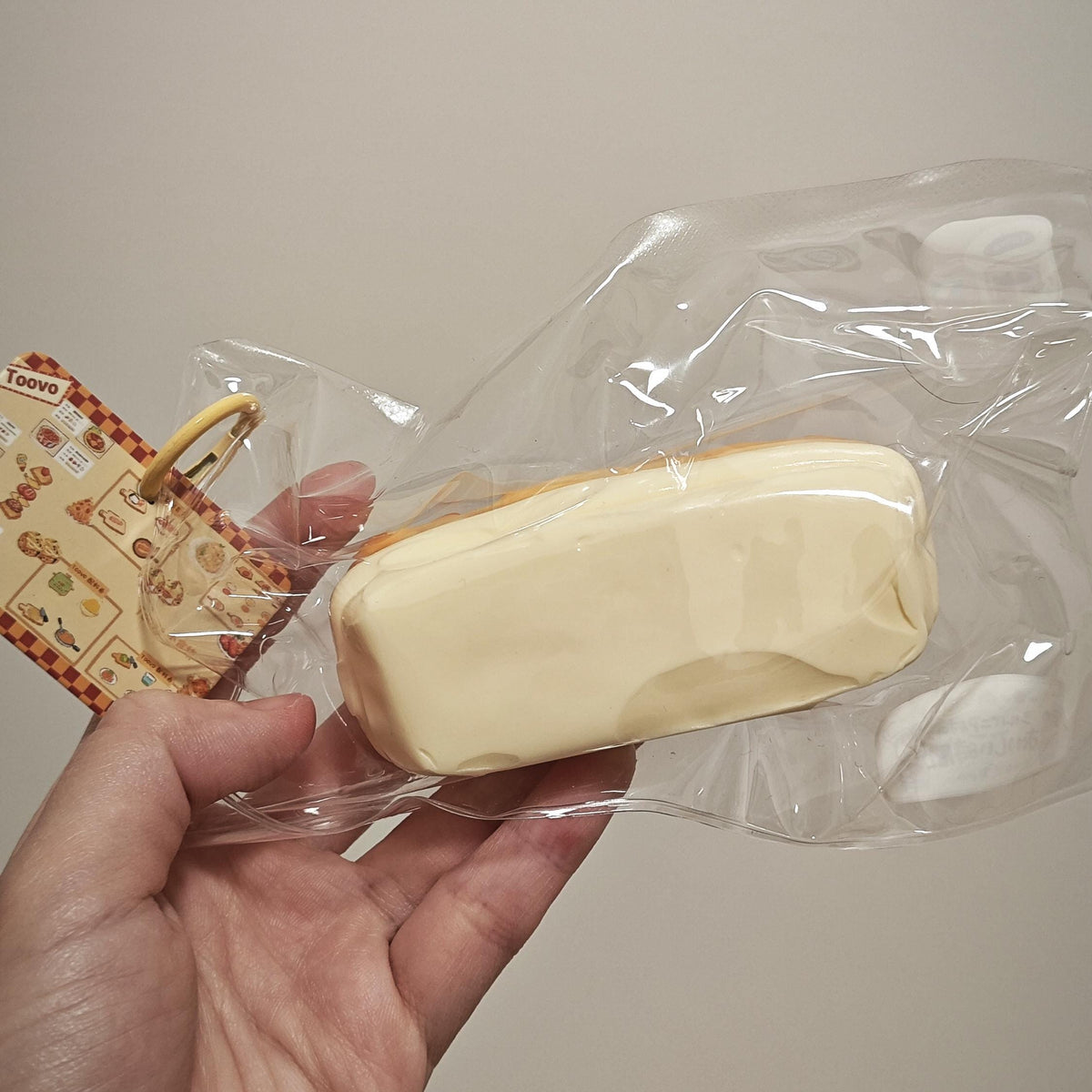 A hand holding a small, rectangular, cream-colored novelty item inside a clear plastic bag with a patterned paper tag attached, against a plain beige background.