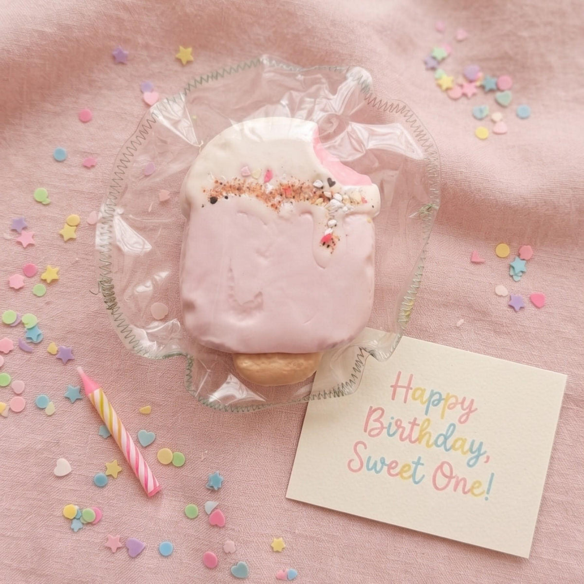 A pink frosted birthday cake topper or decorative cake piece wrapped in clear cellophane, placed on a pink fabric surface with scattered pastel confetti, a small striped candle, and a card that reads 'Happy Birthday, Sweet One!'.
