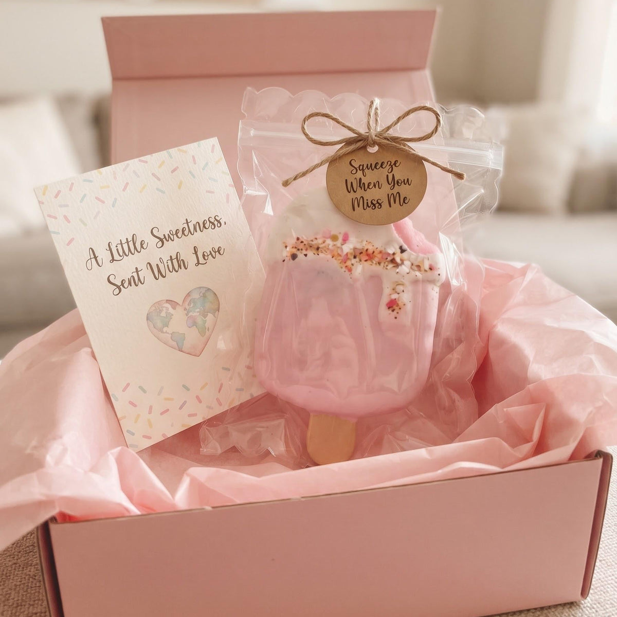 A pink gift box filled with pink tissue paper holds a small wrapped pink item tied with a jute bow and a round tag reading 'Soothe When You Miss Me,' alongside a card that says 'A Little Sweetness Sent With Love.'.