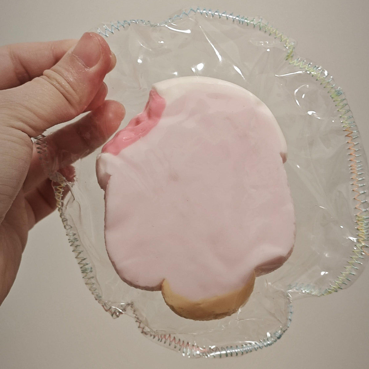 A hand holding a pink frosted cookie inside a clear plastic bag against a plain beige background.