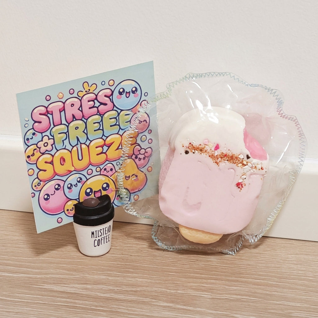 A pink and white squishy toy in clear plastic packaging next to a small jar labeled 'MISTYMOON CRAFTS' and a card that reads 'STRESS FREE SQUEEZ' on a light wood floor against a white wall.