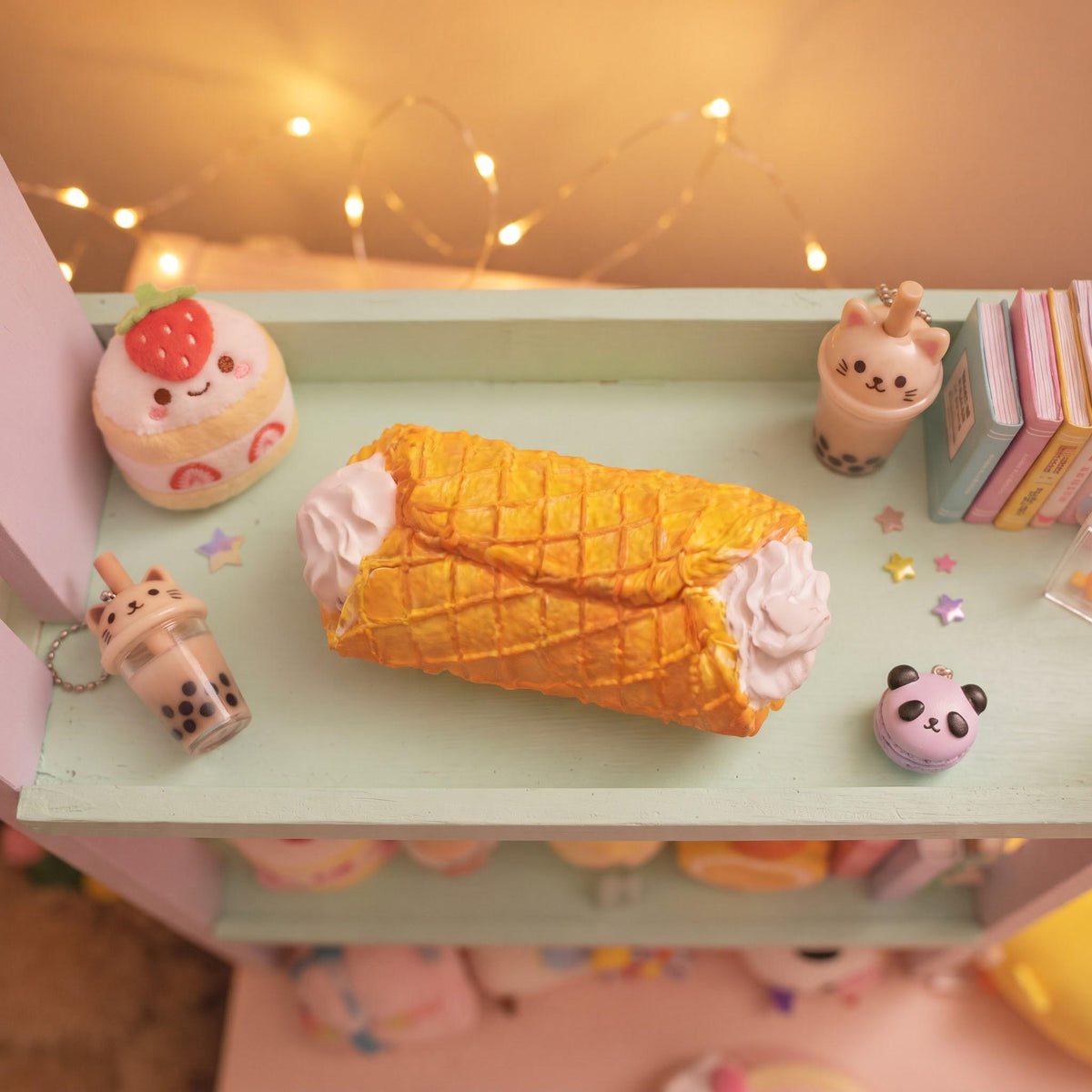 A cute, pastel-colored miniature shelf display featuring a central yellow waffle-patterned pastry with white cream sides, surrounded by small toy desserts, books, and star decorations under warm fairy lights.