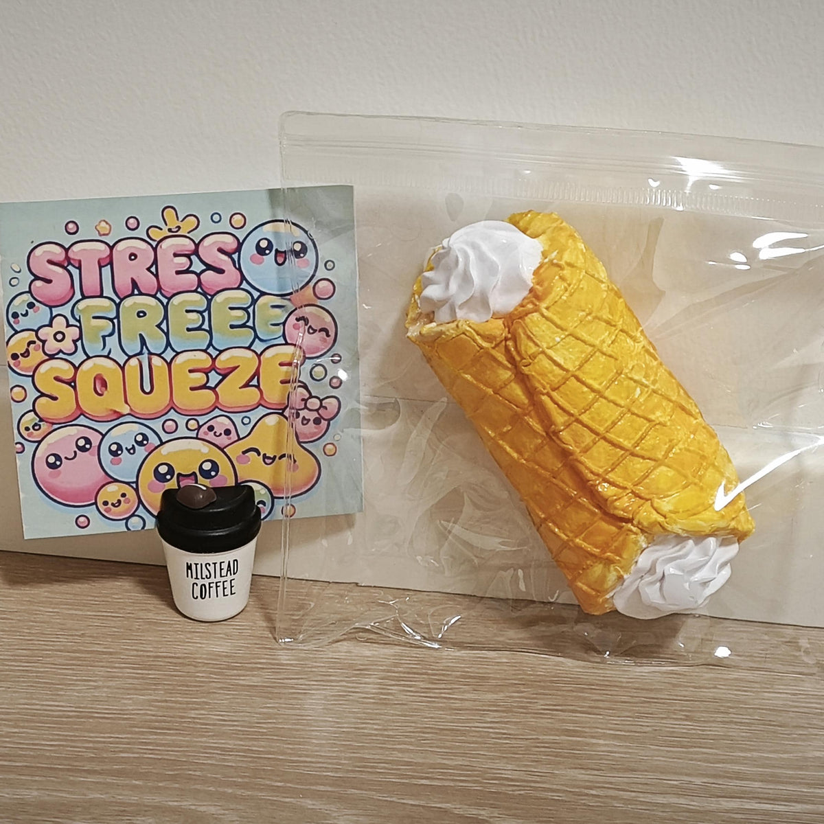 A yellow waffle-cone-shaped squishy toy with white whipped cream ends, sealed in a clear plastic bag next to a square card that reads 'STRESS FREE SQUEEZE' and a small black coffee cup accessory labeled 'Mustard Coffee' on a light wood surface against a beige wall.