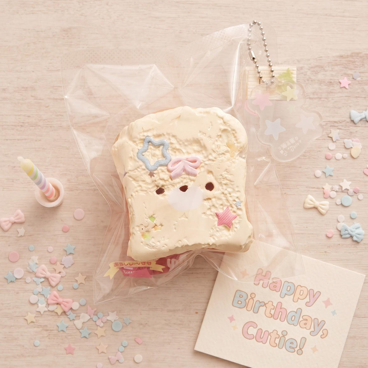 A cute, bread-shaped squishy toy with a smiling face, decorated with pastel star and bow details, packaged in a clear plastic bag on a light wood background with a 'Happy Birthday, Cutie!' card and pastel confetti.