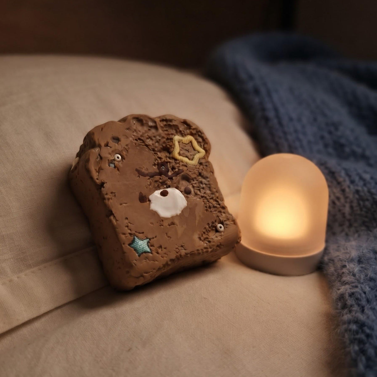 A small decorative object shaped like a brown marshmallow or cookie with a cute face, placed on a bed next to a glowing dome-shaped lamp and a blue blanket.