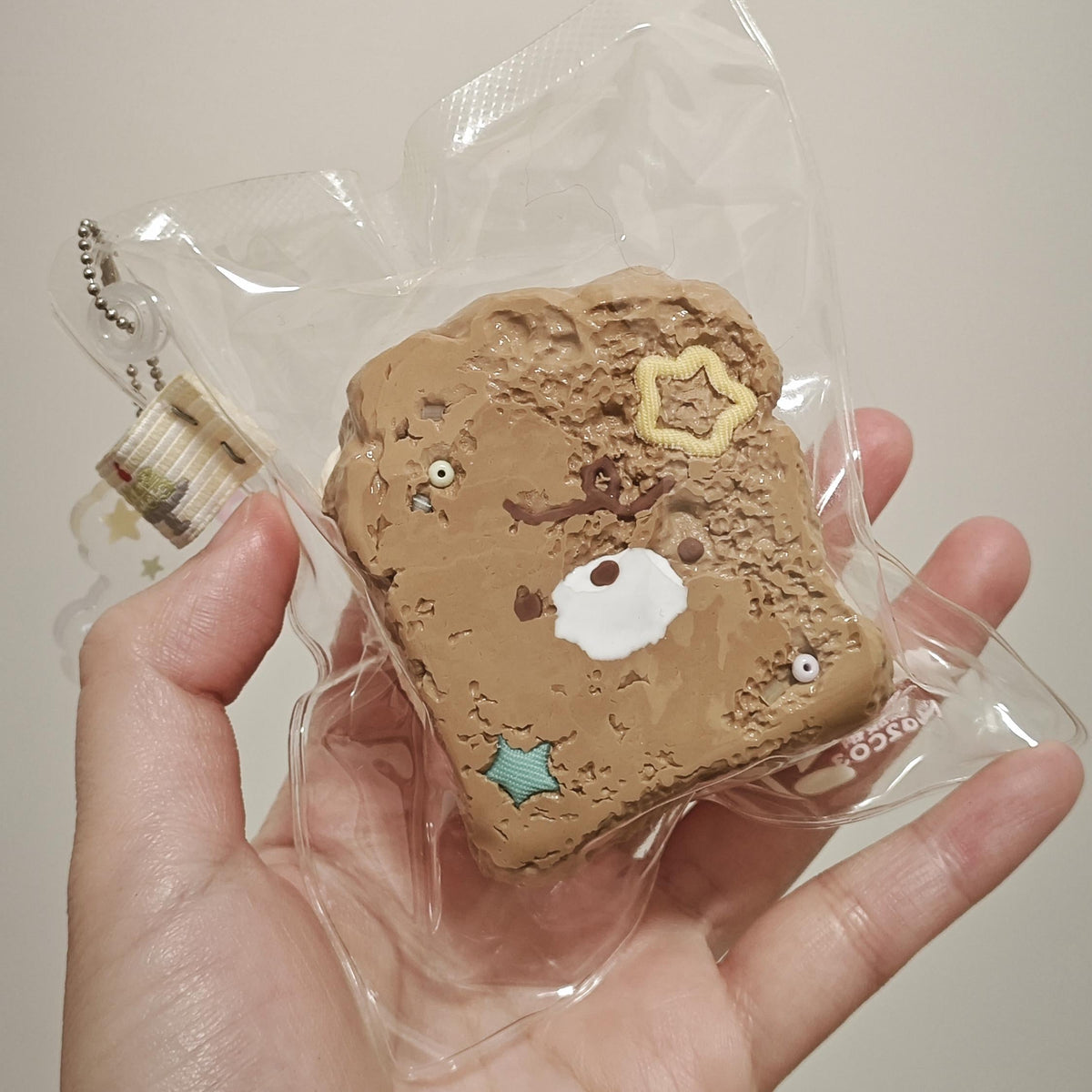 A hand holding a packaged brown bread-slice-shaped squishy toy with cute cartoon face details, sealed in a clear plastic bag against a plain light background.
